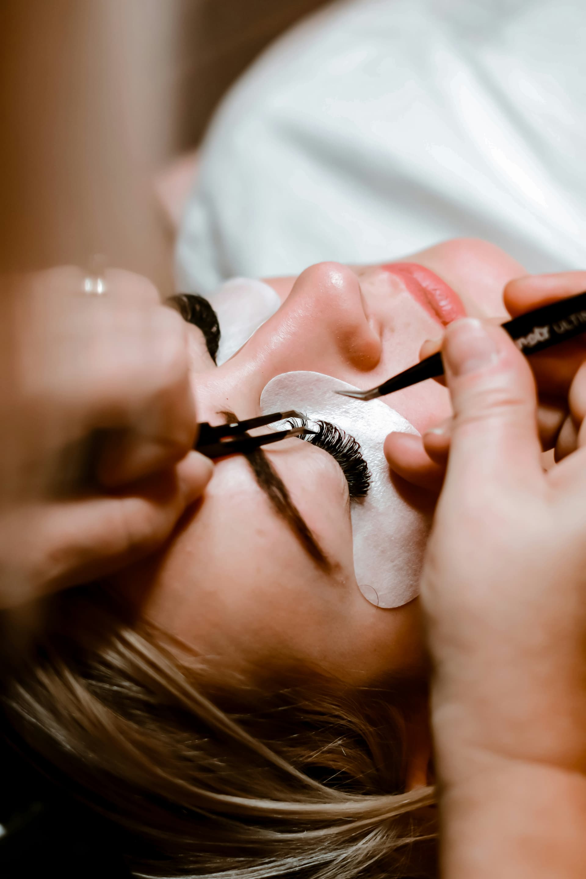 Esthetician carefully applying individual eyelash extensions to client using tweezers and protective eye pad